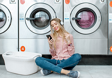 Young woman waiting for the clothes to be washed sitting on the floor and listening to the music at the self-service laundry